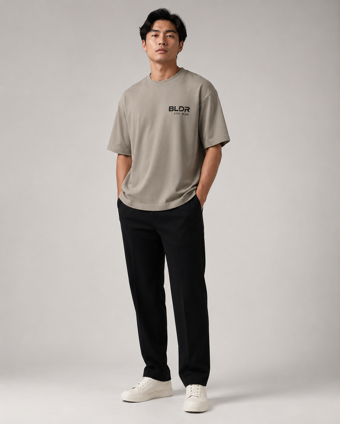 Man wearing a beige t-shirt with a logo, black pants, and white shoes on a gray background